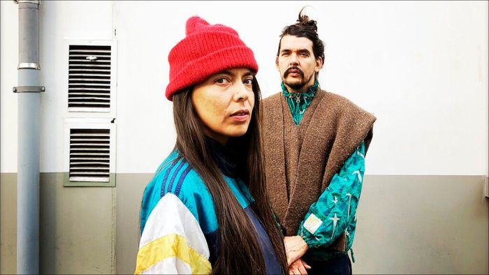 Deux personnes, une femme et un homme, font face à la caméra et portes des vêtements colorés - la femme porte un bonnet rouge. Ils sont face à un mur plutôt industriel / urbain, avec des bouches d'aération et un tuyau de descente (gouttière).