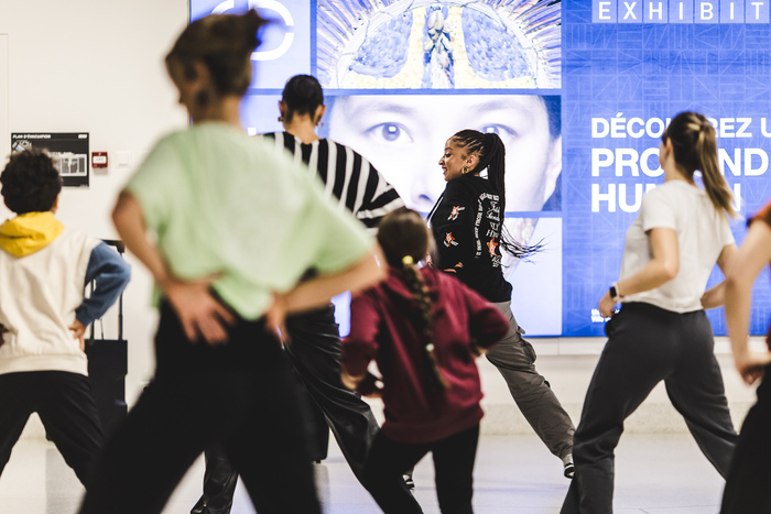DR Photo d'un cours de danse au MEG. La professeurs et de côté et les participant-e-s de dos. Il y a des enfants et des adultes qui participent.
