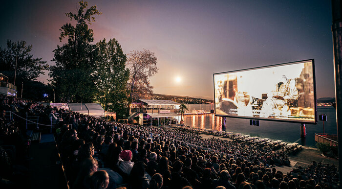 PPR Media Relations AG Un grand écran de cinéma en plein air est installé au bord du lac Léman, avec une foule de spectateurs assis en gradins. La projection a lieu à la tombée de la nuit, sous un ciel violet orangé avec la lune brillante. En arrière-plan, on aperçoit le lac, des arbres et un bâtiment éclairé. L’ambiance est calme et magique.
