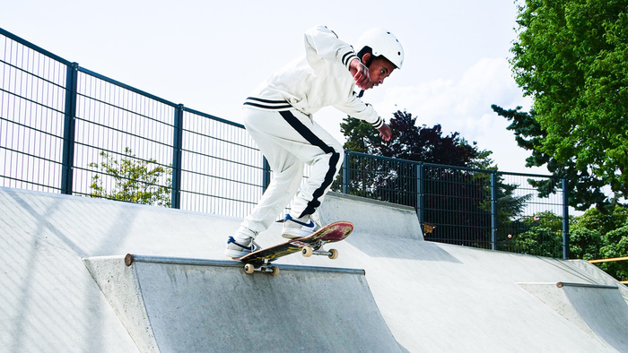 ©Rookie Slash Un jeune skateur habillé en blanc. Il porte un casque blanc et effectue une figure sur une rampe en béton dans un skatepark en plein air.