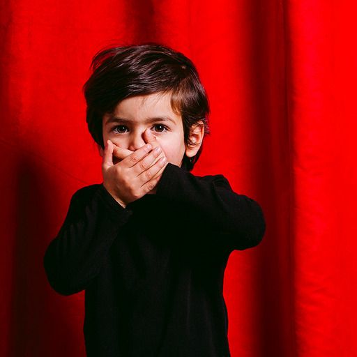 Un enfant se met les mains devant la bouche l'air surpris. Il se tient devant un rideau de théâtre rouge fermé
