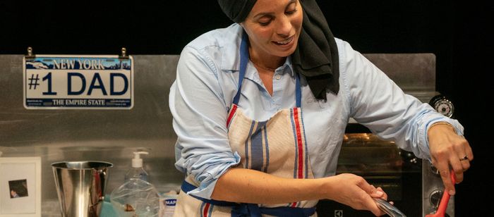 La photographie montre une personne derrière un plan de travail de cuisine installé sur une scène ou dans un espace de spectacle. La personne porte une chemise bleu clair, un tablier à rayures verticales bleu, blanc et rouge, ainsi qu’un tissu noir couvrant la tête et tombant sur une épaule.
Au premier plan, la personne utilise une spatule rouge au-dessus d’un récipient ou d’une préparation situé hors du cadre inférieur. Les mains sont occupées à une activité culinaire.
Derrière le plan de…