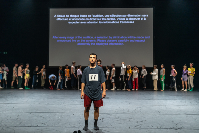 © Gregory Batardon Au premier plan, un danseur avec le numéro 11 sur son t-shirt, en attente, fait face au public. En arrière plan, une ligne de danseurs et de danseuses se préparant devant un écran sur lequel est projeté le texte suivant : A l'issue de chaque étape de l'audition, une sélection par élimination sera effectuée et annoncée en direct sur les écrans. Veillez à observer et à respecter avec attention les informations transmises.