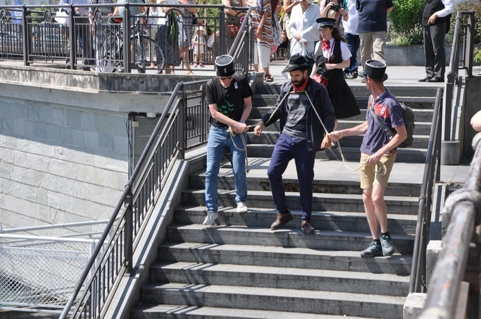 Un comédien de la troupe des Balades Théâtralisées jouant le personnage de l'anarchiste Lucheni, assassin de l'impératrice Sissi, descend des marches. Il est enchaîné et encadré par deux spectateurs coiffés d'un képi de gendarme. La guide et les spectateurs les suivent.
