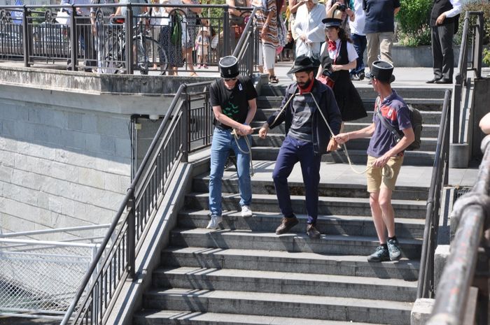 Un comédien de la troupe des Balades Théâtralisées jouant le personnage de l'anarchiste Lucheni, assassin de l'impératrice Sissi, descend des marches. Il est enchaîné et encadré par deux spectateurs coiffés d'un képi de gendarme. La guide et les spectateurs les suivent.