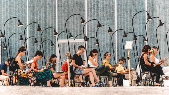 Un groupe de gens est assis sur des chaises sous des lampes - en extérieur - et chacun lit seul quelque chose devant lui.