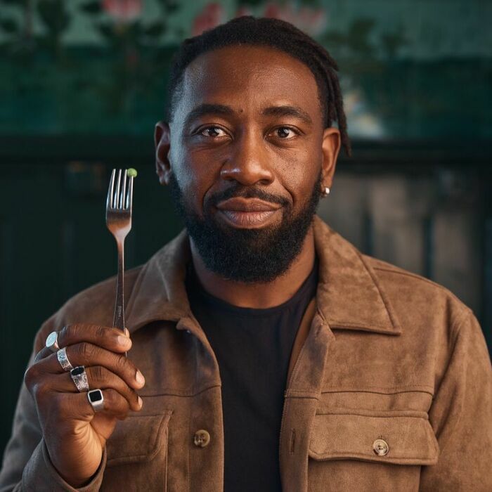 L'humoriste Christian Mukuna, nous regarde, il est habillé d'un veste couleur marron, tient verticalement dans sa main droite une fourchette à hauteur de son visage, avec planté au bout un unique petit pois.