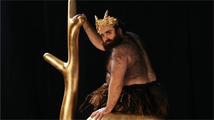 Un homme barbu portant une couronne dorée sur la tête et un pagne tient une (fausse) branche d'arbre dorée.