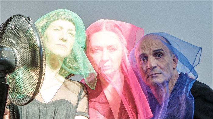 Deux femmes et un homme sont sous des voiles colorés, l'un vert, l'autre rouge et le dernier bleu. Ils regardent la caméra et un ventilateur fait bouger les voiles sur la gauche de l'image.