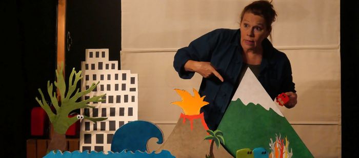 Une personne se tient derrière une maquette colorée représentant un paysage miniature. La personne porte une veste bleu foncé et tient dans une main un petit cube rouge. L’autre main est dirigée vers le décor, comme pour désigner un élément de la scène.
Au premier plan, plusieurs éléments découpés et peints sont disposés sur une table : à gauche, un arbre vert stylisé et un immeuble blanc à fenêtres noires ; au centre, une vague bleue et un volcan gris en éruption, dont le sommet laisse jaillir…