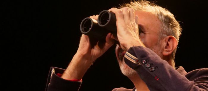 Gros plan d’une personne sur une scène sombre, éclairée par une lumière chaude. La personne porte une veste foncée avec des boutons visibles aux poignets et tient une paire de jumelles noires devant les yeux à l’aide de ses deux mains.
Le cadrage est serré sur le haut du corps et le visage est en grande partie masqué par les jumelles. On distingue des cheveux courts et une barbe grisonnante. Les bras sont relevés pour maintenir les jumelles en position d’observation.
L’arrière-plan est…