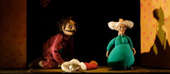 La photographie montre une scène de théâtre de marionnettes éclairée par une lumière chaude. Au centre, sur un plateau en bois, se trouvent deux marionnettes de taille moyenne.
À gauche, une marionnette porte un vêtement rouge foncé à rayures. La marionnette est agenouillée au sol et porte de grandes lunettes rondes. À droite, une seconde marionnette est vêtue d’un costume vert turquoise volumineux et possède de très grandes oreilles blanches qui s’étendent de chaque côté de la tête.
Entre les…
