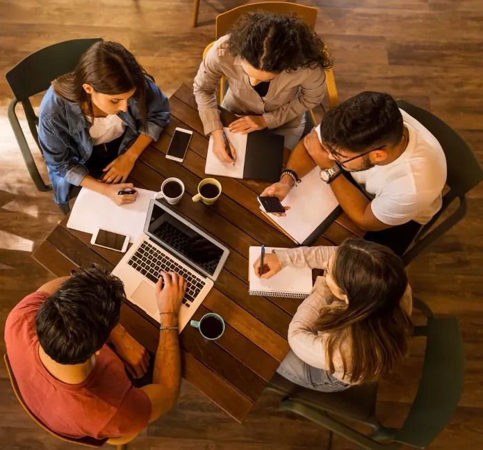 Vue aérienne en plongée de cinq participants à un atelier d'écriture, réunis autour d'une table carrée en bois chaleureux. Dans une ambiance de travail collaborative et conviviale, trois femmes et un homme écrivent à la main avec concentration dans des cahiers ou sur des feuilles blanches, tandis qu'un homme utilise un ordinateur portable. Quelques tasses de café et des téléphones portables sont disposés sur la table, illustrant un moment de création littéraire partagé.