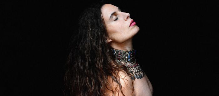 Portrait en gros plan d’une personne photographiée de profil sur un fond entièrement noir. La tête est légèrement inclinée vers l’arrière et les yeux sont fermés. Les cheveux longs et ondulés tombent sur les épaules.
La personne porte un imposant collier métallique couvrant largement le cou et le haut de la poitrine. Ce bijou est composé de multiples rangées d’éléments argentés ornés de petites pierres colorées, avec plusieurs pendentifs suspendus à l’avant.
L’éclairage met en valeur le visage,…