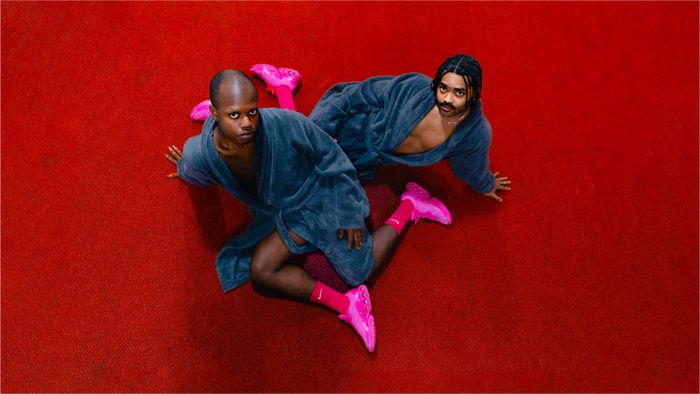 Deux hommes en peignoir bleu avec des baskets roses regardent la caméra. Ils sont allongés sur un tapis rouge.
