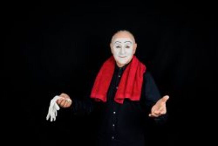Un homme écartant les bras. Il est habillé de noir. Il a des gants blancs dans une main et une serviette rouge sur les épaules. Un masque blanc est dessiné sur son visage