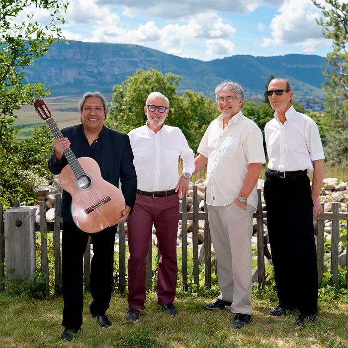 Quatre hommes souriants debout dont un avec une guitare en main