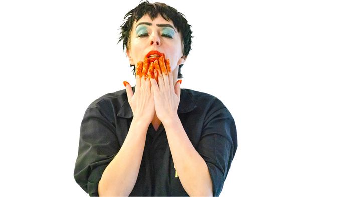 Une femme met ses mains à son visage, elles sont maculées de peinture orange. La femme est sur un fond blanc avec une blouse noire.