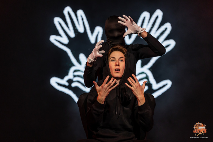 Deux personnes (une femme bien visible et un homme avec un masque noir) montrant leurs mains et il y a quatre mains lumineuses derrière eux