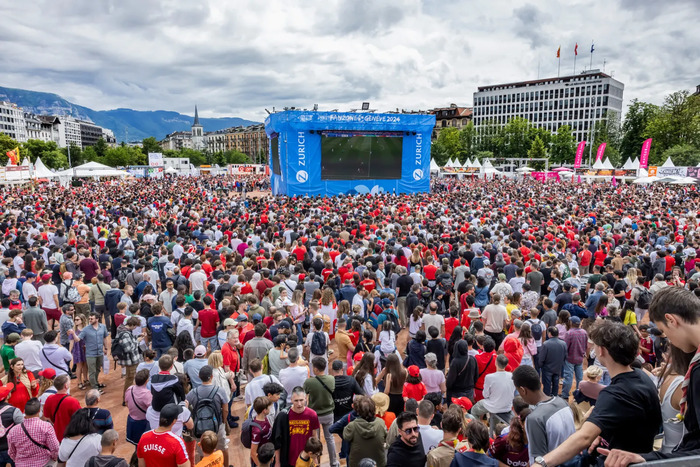 DR Une foule dense de milliers de personnes, majoritairement vêtues de rouge, est rassemblée en plein air devant un grand écran bleu marqué « Fan Zone Genève 2024 ». L’ambiance est festive. Des tentes blanches bordent les côtés, et des montagnes sont visibles en arrière-plan sous un ciel nuageux. On aperçoit aussi des drapeaux, des bâtiments et une église au loin.