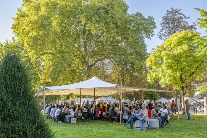 L'image montre une grande tente blanche, installée sur une vaste pelouse verte. Sous cette tente ouverte, de nombreuses personnes sont assises sur des bancs autour de longues tables en bois, partageant un repas.