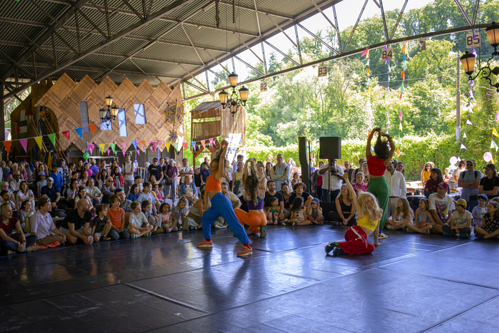 Quatre danseuses se tiennent sur une scène au premier plan. En second plan, on distingue le public assis, composé de nombreuses personnes. La scène se situe dans un hangar ouvert, rappelant une ancienne structure industrielle. On observe que le lieu a été transformé à des fins socio-culturelles, avec la présence de petites constructions.