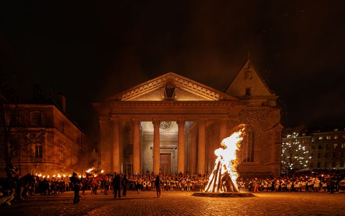 Un grand feu de joie brûle sur sur le parvis de la Cathédrale Saint-Pierre. Une foule nombreuse, dont beaucoup de porteurs de flambeaux et de torches, entoure le brasier dans la nuit. On distingue aussi le Héraut et son cheval au premier plan. La scène dégage une ambiance très solennelle et festive, typique d'une commémoration nocturne comme celle de l'Escalade à Genève.