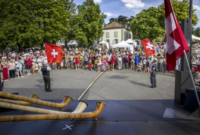 Magali Girardin / Ville de Genève L'image est une photographie d’une célébration de la Fête nationale suisse à Genève en plein air, par une journée ensoleillée. Au premier plan, plusieurs cors des Alpes, longs instruments en bois, sont posés devant une scène. Deux hommes en costume traditionnel brandissent de grands drapeaux suisses rouges avec la croix blanche, face à une large foule rassemblée en demi-cercle. Les spectateurs, debout, comprennent des familles, des enfants et des personnes de tous âges. En arrière-plan, on…