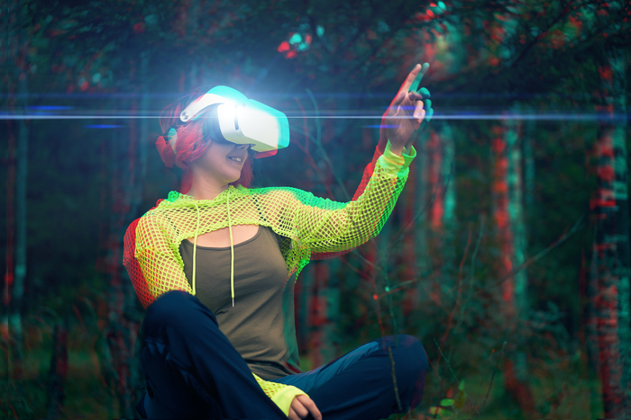 Une femme assise dans l'herbe, habits futuristes ou sport technique, un casque VR sur les yeux, lève le doigt comme pour toucher quelque chose devant elle. C'est dans la forêt, l'image est glitchée, comme décalée en rouge et bleu.