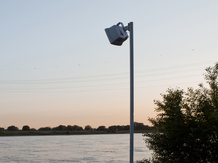 Sur cette image on voit un haut-parleur fixé en haut d’un mât métallique, installé au bord d’un fleuve.
