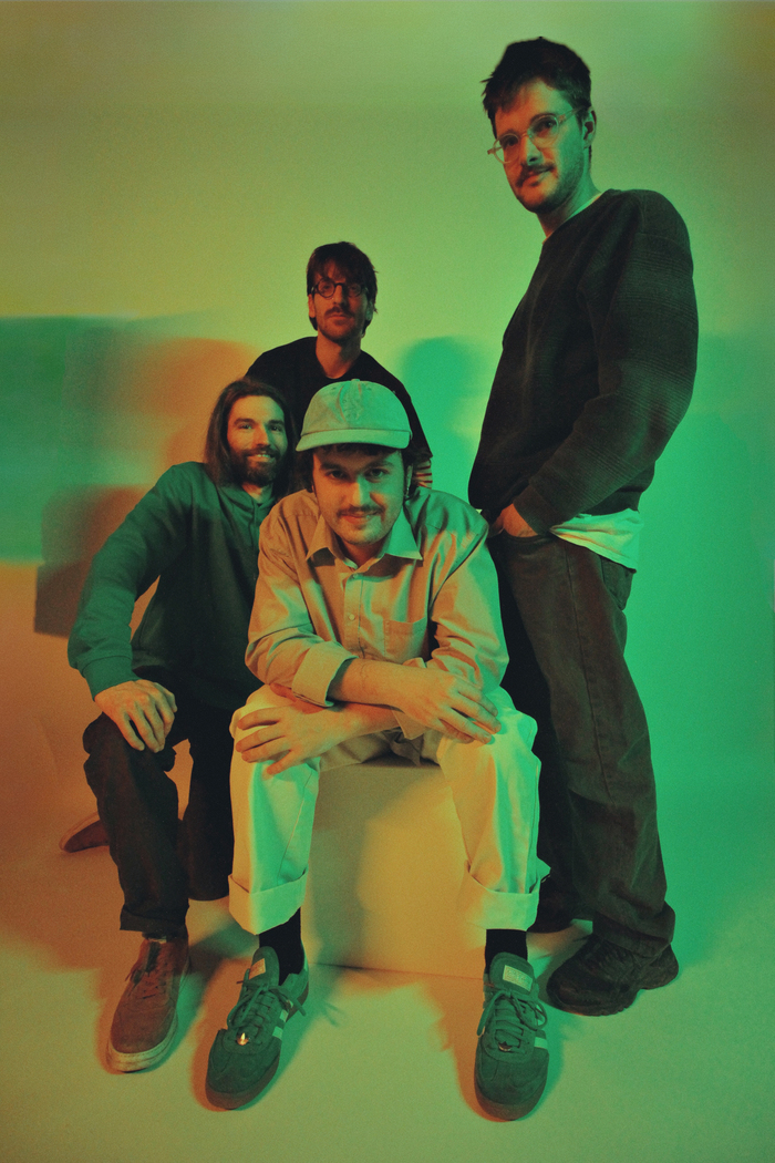 Photo promotionnelle de l’événement Jazztronica #3. Quatre musiciens posent dans un studio éclairé de lumières vertes et orangées. Un membre est assis au centre, casquette claire et chemise beige, les trois autres se tiennent debout derrière lui, souriants, en tenues décontractées.