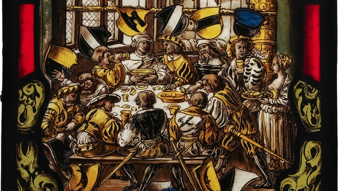 © Atelier Girardet Vitrail représentant un festin médiéval : on y voit des hommes autour du table en train de manger et de boire. Il y a une dominance de jaune et de noir.