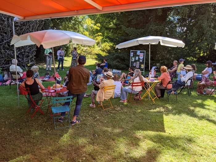 Paola Emery Tables rondes et chaises avec des parasols disposées dans le jardin. Des gens écoutant des orateurs
