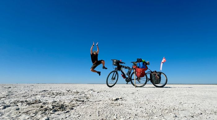 Un homme saute de joie dans un désert blanc, à côté de son vélo. Le ciel est d'un bleu intense en arrière-fond.