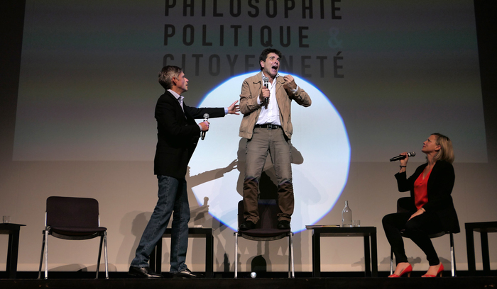 Carole Favre Un intervenant très engagé debout sur une chaise face à l'audience parle dans un micro et deux de ses collègues essaient de le calmer.