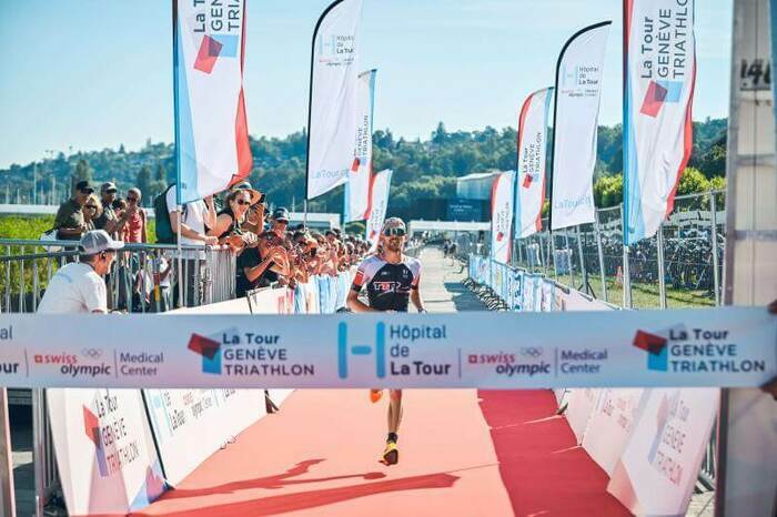 DR Sur l'image, on voit l'arrivée du Tour Genève Triathlon avec un coureur arrivant seul. De chaque côté, une foule l'encourage et des drapeaux portant le nom de l'événement sont visibles. Un tapis rouge est déposé sur le sol à l'arrivée. Le ciel est bleu, et en arrière-plan, on aperçoit des arbres verts.