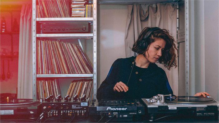 Une femme mix sur des platines de DJ dos à une étagère recouverte de CDs et vinyles.