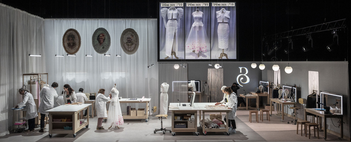 Sur cette image, nous pouvons voir plusieurs personnes travaillant dans un atelier de couture sur différents modèles et à différents endroits de la pièce. Il y a également en second plan sur un écran l'image de 3 robes.