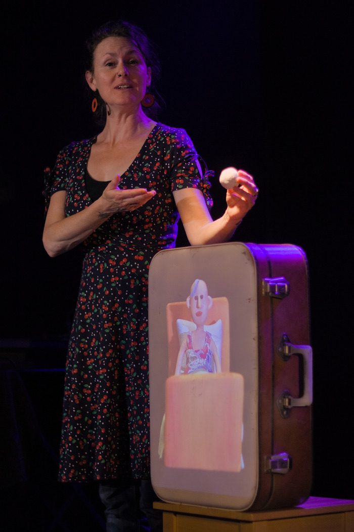 Jeune femme debout à côté d'une valise où est projetée l'image d'une femme couchée. La femme couchée est un personnage en pâte  à modeler