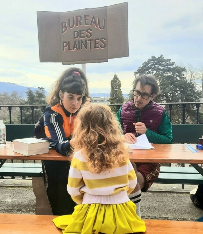 Cie Sale Bête production Deux adultes assis derrière une table regardent une enfant, dos à nous, de l'autre côté de la table. L'homme a un micro. Derrière eux, écriteau "Bureau des plaintes"