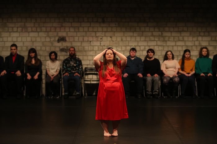 Sur cette image, il y a en premier plan une femme seule, vêtue d'une robe rouge se tenant la tête entre les mains. Derrière elle au second plan il y a neuf personnes assises qui l'observent.