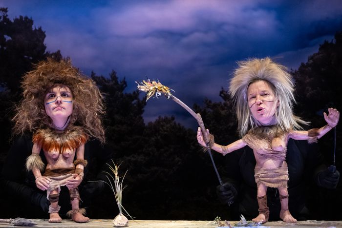 Deux personnages préhistoriques féminins en marionnettes Kokoschka font face au public. L'une d'entre-elles tient dans sa main un bâton en feu.