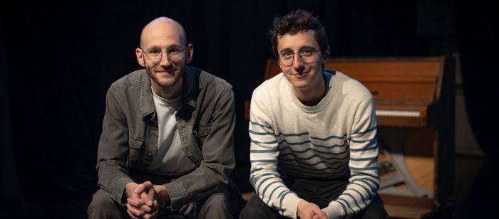 Photographie prise sur une scène de théâtre ou dans un espace de spectacle faiblement éclairé. Deux personnes sont assises côte à côte au premier plan, face à l’objectif.
La personne située à gauche porte des lunettes rondes, un t-shirt clair sous une veste grise et un pantalon assorti. La tête est rasée. Les mains sont jointes devant les genoux.
La personne située à droite porte également des lunettes rondes, un pull blanc à rayures horizontales bleues et un pantalon foncé. Les avant-bras…