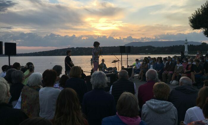 Un groupe de personnes est rassemblé en plein air au bord d’un lac, face à une petite scène. Un artiste parle au micro devant un public attentif, assis ou debout. Le soleil se couche à l’horizon, baignant le ciel de couleurs chaudes. L’ambiance est paisible et conviviale, avec vue sur l’eau, les montagnes et un phare blanc.