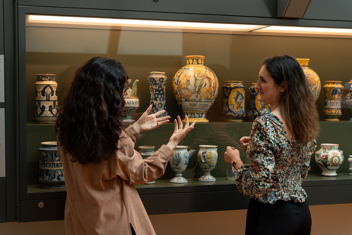 Une médiatrice donne des informations à une visiteuse sur une vitrine présentant des pots de pharmacie.