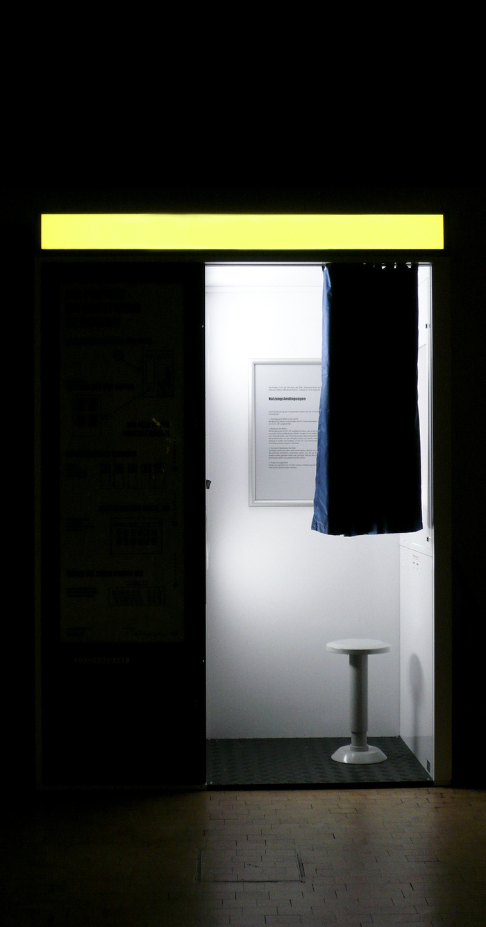 © Magali Dougados C'est l'image d'un photomaton dans le noir. Il y a de la lumière dans la cabine on aperçoit un petit rideau et un petit tabouret.