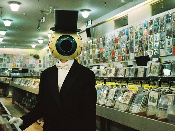 Sur cette image, on voit une personne avec un gros œil à la place de la tête. Elle porte un costume noir et fixe la caméra, dans un magasin de disques