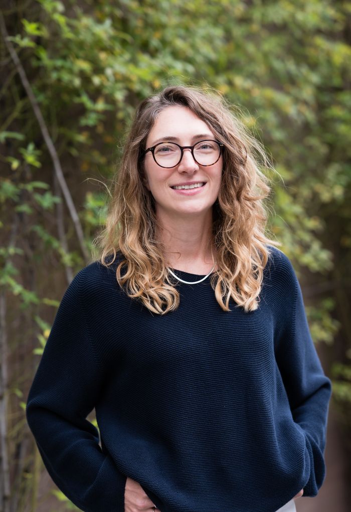 Portrait de l'intervenante jusqu'à la taille. Cheveux longs plutôt bouclés clair. Elle a un pull bleu foncé les mains dans les poches, des lunettes rondes et elle sourit.