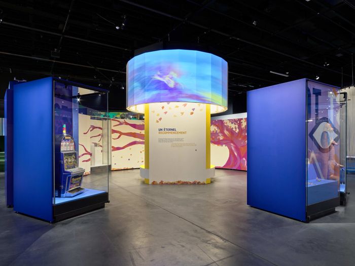 Prise de vue de l'exposition Le futur c'est quoi, on y voit au centre une projection en forme de rond sur une structure droite et sur les côtés des vitrines avec des objets.