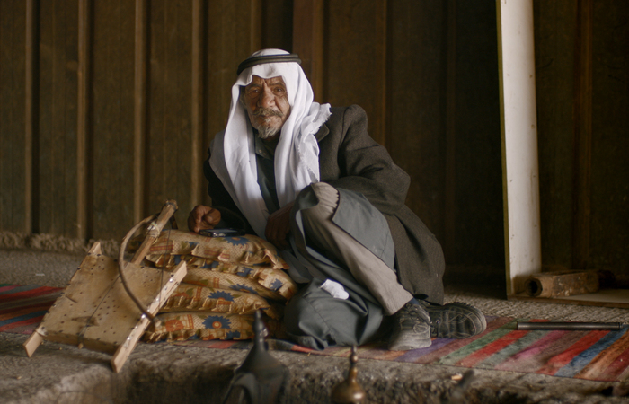 vieil homme, tenue traditionnelle, keffieh blanc (Foulard du Moyen-Orient), regard pensif, intérieur sombre, tapis coloré, coussins à motifs, instrument à cordes, artisanat, ambiance rustique, vêtements amples, bâton, lumière naturelle, regard